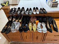 Overview of 17 pairs of assorted women's shoes lined up on and in front of a wooden dresser, displaying various styles and colors, some wrapped in plastic bags.