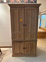 Front view of wooden wardrobe or entertainment center showing two large double doors on top and two large double doors below with black knobs, natural wood finish and crown molding on top.