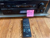 Front view of Yamaha RX-V2300 AV receiver with remote placed in front on hardwood floor. Receiver display visible with text and controls on front panel.