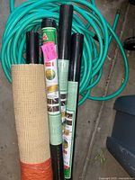 View showing coiled green garden hose, beige woven yard fabric rolled up, three black landscaping fabric rolls beside hose