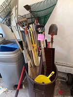 Photo showing various yard tools including shovels, rakes, a garden hoe, small hand tools, and a brown plastic planter pot arranged in a corner by wall and storage bins.