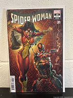 Photo of Spider-Woman #16 comic book variant cover featuring Spider-Woman and Deadpool in costume, sealed in protective sleeve, showing cover art and issue number.