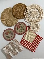 Overview of needlepoint floral coasters, wicker trivets, red and white pot holder, and beige dishcloth