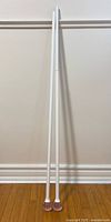 Full view of two white adjustable curtain rods leaning against a wall showing their length and pink bulbous ends.