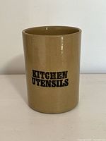Front facing photo of a beige glazed stoneware crock with black text 'KITCHEN UTENSILS' printed clearly in two lines.