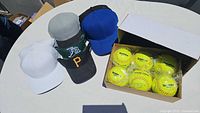Group of five hats and visors displayed on white table including white, gray, blue caps and two visors, next to box of six yellow Richardson softballs.