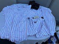 Group of nine white Wilson youth baseball jerseys with red vertical pinstripes arranged flat on a table, with one black and white sleeveless striped jersey on top.