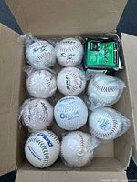 Top-down image showing a box containing 11 softballs, mostly wrapped in plastic, including Dudley Thunder SW, Wilson OPS, and MacGregor Duzz-Ezy (in box).