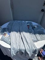 Photo showing men's Wilson baseball pants in grey with black vertical stripes laid on a white table, some pants unpackaged, some still in plastic packaging with tags visible.