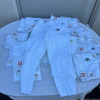 20 white Wilson youth baseball pants displayed on table, mostly in bags with tags visible.