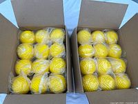 Two open cardboard boxes each containing 12 individually wrapped yellow pitching machine balls with dimpled texture, totaling 24 balls.