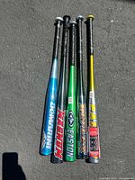 Group of five aluminum baseball bats standing upright on pavement, showing their length and grip taped handles.