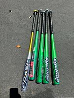 Five aluminum baseball bats laid out side by side on pavement, showing color and grip condition.