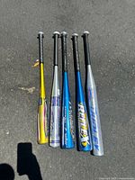 Group view of 5 aluminum baseball bats laid side by side on pavement showing colors and grips