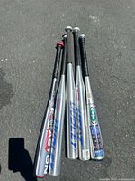 Five aluminum baseball bats laid side by side on pavement, showing barrels and grips.