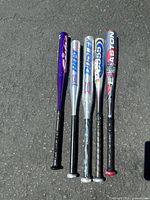 Photo showing five aluminum baseball bats laid on pavement including brands Easton, Louisville Slugger, and Dudley. Variations in color and design visible.