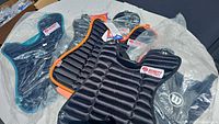 Five new catcher's chest protector pads arranged on a table, showing various colors including black, teal, and orange, with visible packaging and brand labels.
