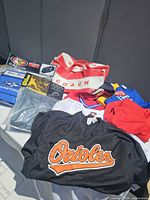 Photo of the group of various sports items including black Orioles shirt, red and white Coach bag, and multiple other sports shirts and bags.