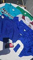 Various sports and casual tops laid out on table including a teal MARIN jersey, blue mesh jersey with number 0, green T-shirt, and blue, white, maroon pirate logo top.
