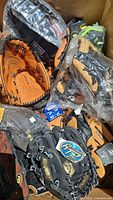 Box filled with various youth leather baseball mitts, some individually wrapped, showing different brands and colors.