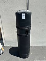 Black tackling dummy standing upright against a plain light wall, showing full cylindrical shape and two side handles with straps.
