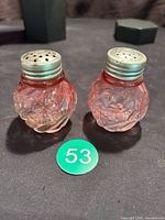 Pair of cranberry glass salt and pepper shakers with metal tops featuring leaf design and umbrella shape, shown on black surface with green number 53 token.