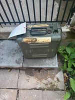 Front-facing view of the olive drab green plastic carrying case with handle, showing wear, old military labels, and the lockable latch hardware on top.