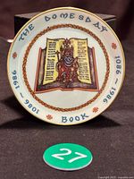 Top-down photo of the commemorative plate showing detailed artwork of seated figure on open book with text and gold rim, highlighting the inscription 'THE DOOMSDAY BOOK 1086 - 1986'