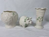 Three white ceramic Belleek pieces: Celtic mini cache pot with shamrocks, piggy bank with shamrocks, and vase with embossed patterns and scalloped rim.