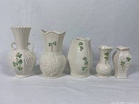 Front view of five Belleek vases arranged side-by-side, showing varying shapes, embossed textures, and shamrock decorations.