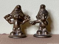 Two brass Smurf figurines side by side on a flat surface, showing poses and wear
