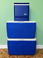 Three coolers stacked against green wall: two blue Coleman coolers with white lids (24x14") stacked on bottom, blue Igloo Legend cooler (13x24") on top.