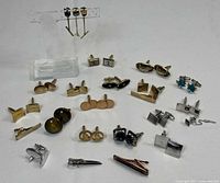 Wide view showing full assortment of mid century style cuff links and tie pins on a white background, including various metal finishes and some colored stone accents.
