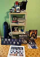 Photo of shelving unit holding mugs, glasses, books, and other memorabilia with additional items displayed on floor including framed images and decorative plaques.