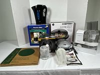 Full lot displayed including Proctor Silex coffee maker in box, Pampered Chef round stone, Chefman electric skillet in box, Keurig thermal carafe in box, white food processor, coffee filters, pods, and measuring cups.