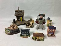 Overall view showing eight miniature house figurines arranged on a white background. Includes rustic log cabin, stone castle, white house, stone tower, yellow building, brick multi-story building, and two thatched roof cottages.