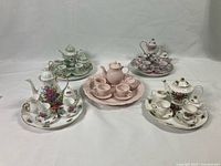 Photo of five miniature porcelain tea sets arranged on a plain white background, showing various flower and gold-accent designs, with cups, saucers, teapots, sugar bowls, and creamers visible.
