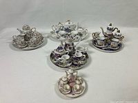Five assorted miniature porcelain tea sets arranged on trays with teapots, cups, saucers, creamers, and sugar bowls.