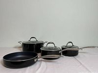 Photo of four black cookware pieces with stainless steel handles and glass lids, showing an overview of the set.