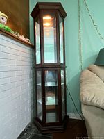 Full view of cherry finished wooden curio cabinet placed between wall and couch, showing light on and glass shelves.