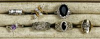 Top view of 8 assorted rings arranged on beige display: various stones, metals and designs visible