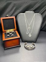 All items displayed together on black background: rings in wooden box, necklaces on bust, bracelet and earrings front