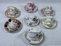 Overhead view showing all seven assorted teacup and matching saucer sets with floral and decorative patterns.