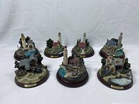 Photo of seven Thomas Kinkade Guiding Light cottage and lighthouse collectible figurines arranged in two rows on a white background, showing detailed designs of each piece.
