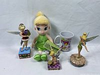 Front view of Tinker Bell plush, mug, and three Disney Traditions figurines together on a white background