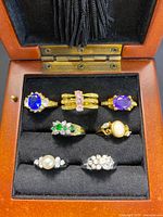 Wooden ring box containing eight vintage rings in gold and silver colors with various colored stones, displayed in velvet slots.