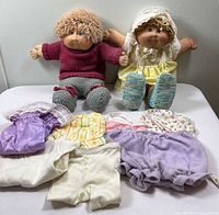 Two vintage Cabbage Patch Kids dolls sitting on a white surface with an assortment of seven doll clothing pieces laid out in front of them.