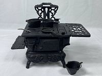 Front angle view of vintage Crescent cast iron miniature stove with pots, pan, ladle on top and bucket beside it