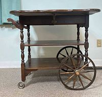 Side view showing the tea cart with large wooden spoked wheels on one side and smaller caster wheels on the other; foldable top partially open