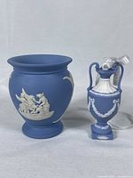Two blue Wedgwood jasperware pieces: a vase and a miniature urn, both with white classical reliefs on a matte blue finish.